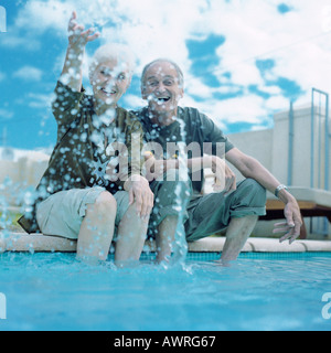 Coppia matura seduta sul bordo della piscina, donna gli spruzzi di acqua nell'aria Foto Stock