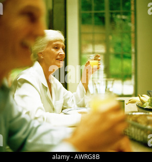 Donna matura seduta a tavola, tenendo il vetro, uomo sfocato in primo piano Foto Stock