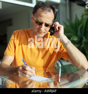 Uomo maturo utilizzando phone, prendendo appunti a tavola Foto Stock