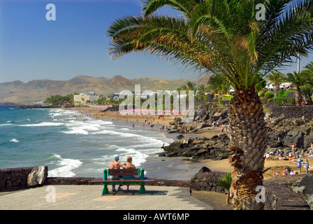 PUERTO DEL CARMEN giovane seduto sulla panchina incorniciata da palme affacciato sulla soleggiata spiaggia sabbiosa a Puerto Del Carmen Lanzarote isole Canarie Spagna Foto Stock