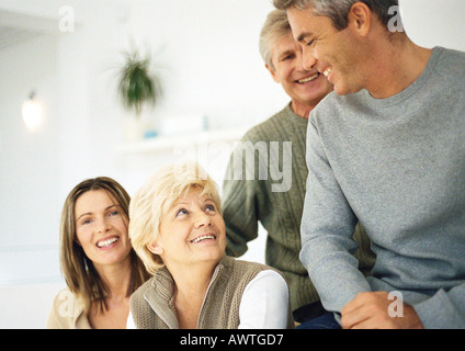 Due donne sorridenti, testa e spalle, uno guardando in alto, due uomini, vita fino in salotto impostazione Foto Stock