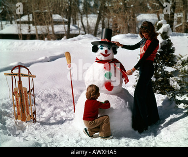 Madre e figlio giovane mettere i tocchi di rifinitura su un jaunty pupazzo di neve in una variopinta scena invernale Foto Stock