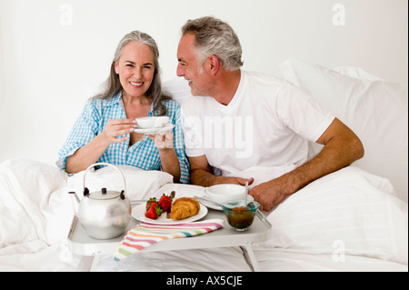 Coppia matura seduta sul letto con colazione, sorridente Foto Stock