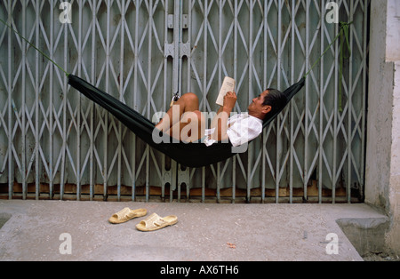 Un uomo si siede in una amaca e legge un libro al di fuori della porta chiusa di un negozio in Vietnam Foto Stock