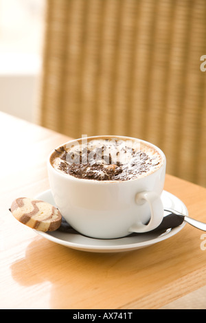cappuccino in tazza bianca e piattino e biscotto con sedia color paglierino dietro. Foto Stock