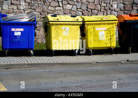 Contenitori di riciclaggio blu, gialli e arancioni per lo smistamento dei rifiuti su un marciapiede pavimentato con un muro di pietra e erba verde sullo sfondo sotto la luce del sole. Foto Stock