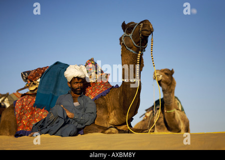 Un ritratto di un cammello conducente con le sue due cammelli nel deserto pomeriggio con la Luna in cielo dietro di loro. Foto Stock