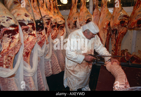 Circondato da appendere le carcasse, una macelleria seghe in corrispondenza di un giunto di carne di carne di Smithfield Market nella città di Londra, Regno Unito. Foto Stock