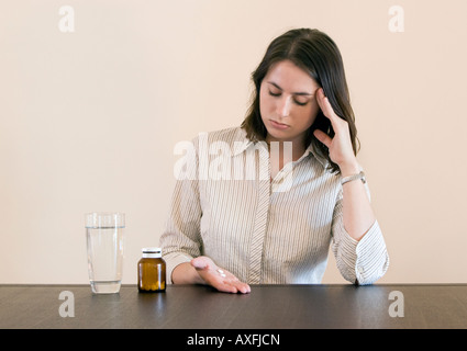 Una giovane donna con un mal di testa si sedette ad un tavolo con due compresse in mano e le dita per i suoi templi Foto Stock