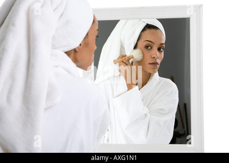 Bella giovane donna messa sul make up di fronte a uno specchio indossando i suoi capelli in un asciugamano e accappatoio bianco Foto Stock