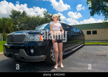 Una giovane donna si appoggia su un nero limousine allungata con un garage, fogliame, cielo blu e bianchi e soffici nuvole in background. Foto Stock