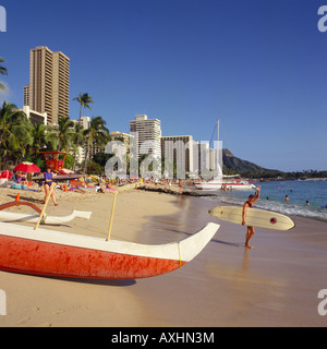 La spiaggia di Waikiki con surfer sventolando la fotocamera che porta la sua tavola da surf al mare nei pressi di canoa outrigger Honolulu Hawaii Foto Stock