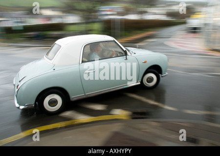 Piccolo due posti in stile retrò pallido color aqua Nissan Figaro auto guidato sotto la pioggia con motion blur per accentuare il movimento Foto Stock