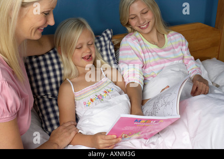 Madre seduta sul letto delle sue due figlie bionda, tutti guardando un libro per bambini, close-up Foto Stock