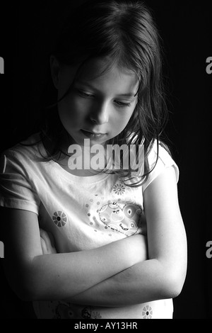 Colpo di un grazioso 7 anno vecchia ragazza guardando in giù con le braccia incrociate, contro uno sfondo nero Foto Stock