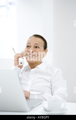 Imprenditrice tramite telefono fisso in ufficio, lo sguardo lontano, sorridente Foto Stock