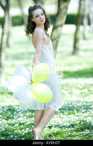 Giovane donna camminare in prato, tenendo palloncini, guardando sopra la spalla in telecamera Foto Stock