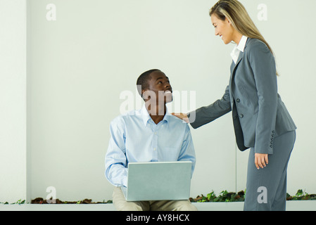 Ragazzo adolescente seduta, utilizzando il computer portatile, sorridente fino a imprenditrice in piedi accanto a lui Foto Stock