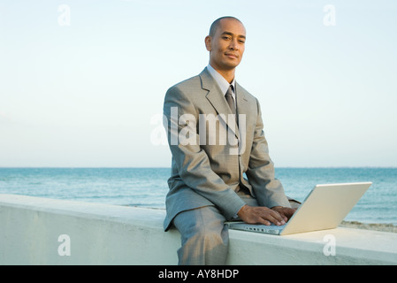 Imprenditore seduto sul muro basso dal mare, utilizzando il computer portatile, sorridente in telecamera Foto Stock