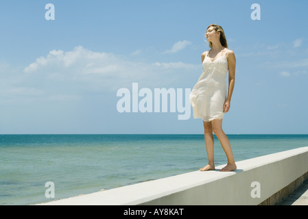 Giovane donna in sundress permanente sulla bassa parete accanto al mare, gli occhi chiusi Foto Stock