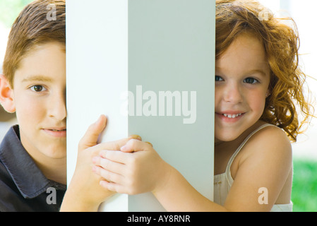 Fratello e Sorella di peeking attorno alla colonna in telecamera, tenendo le mani, sorridente Foto Stock