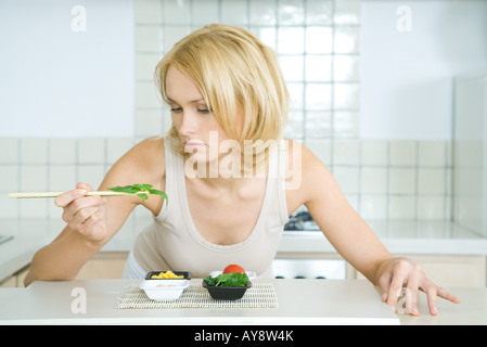 Giovane donna sporgendoti bancone cucina, prelievo di erbe con bacchette Foto Stock