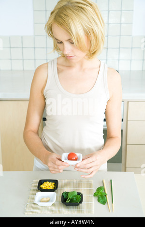 Giovane donna in piedi in cucina, tenendo cherry pomodoro nel piatto piccolo, guardando verso il basso Foto Stock