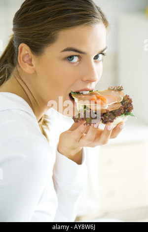 Donna di mordere in sandwich, guardando sopra la spalla in telecamera Foto Stock