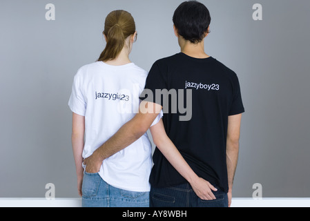 Coppia giovane indossando personalizzato tee-shirt, braccia intorno reciprocamente le cinture, vista posteriore Foto Stock
