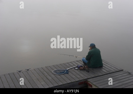Lone uomo crosslegged seduto su di un molo in legno, pesca nella nebbia di mattina. Foto Stock