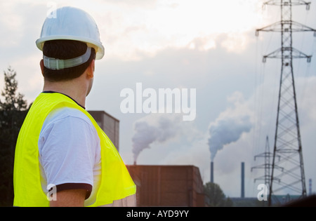 Un uomo vestito di compressori hardhat e di sicurezza ad alta visibilità indagini camicia una fabbrica con camini mettendo fuori il fumo Foto Stock