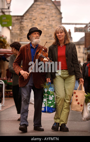Un musicista ELDERLEY suona un violino che accompagnano una signora lo shopping al mercato degli agricoltori in Stroud GLOUCESTERSHIRE REGNO UNITO Foto Stock