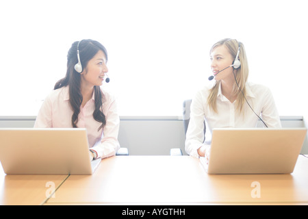 Due imprenditrici che indossa gli auricolari e lavorare con i computer portatili in ufficio Foto Stock