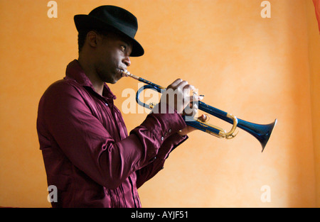 African American uomo che indossa un cappello Fedora riproduce una tromba blu davanti a una parete gialla all'interno Foto Stock