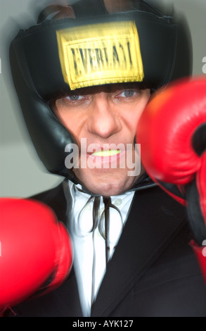un uomo d'affari che indossa una protezione per la testa e un mouthguard solleva i suoi guanti rossi vicino al viso, in posa da boxe Foto Stock