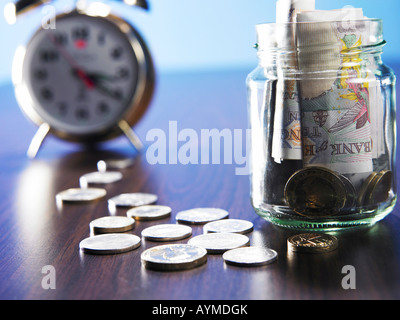 Un vaso pieno di monete con monete e sveglia sul tavolo Foto Stock