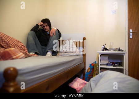 giovane uomo teenage con capelli scuri seduto alla fine del suo letto in camera da letto disordinata arricciato fino al muro esclusione sociale rimanendo a casa Foto Stock