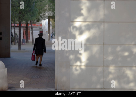 Una signora anziana la sacca di custodia si allontana dalla telecamera in ambiente urbano con venature ombre sui muri in primo piano Foto Stock