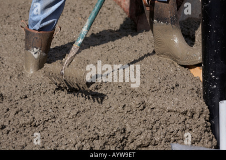 Workman in stivali di protezione e jeans utilizzando un rastrello per appianare i concreti e spostarlo intorno a tubi di accesso durante la posa del calcestruzzo Foto Stock