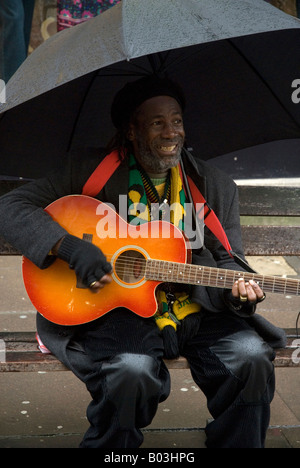 Il giamaicano musicista di strada seduta su una panchina sotto un ombrello a suonare la chitarra, bagno, Somerset, Inghilterra, Regno Unito Foto Stock
