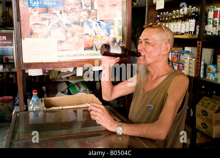 Barbuto Chinaman bere birra da una bottiglia nel suo negozio, Port Louis market, Maurizio Foto Stock