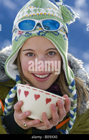 Giovane donna con cappello e occhiali da sole, il riscaldamento se stessa con una tazza di tè caldo, Francia, Alpi Foto Stock