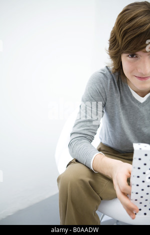 Giovane uomo seduto, tenendo borsa regalo, sorridente alla fotocamera, vista ritagliata Foto Stock