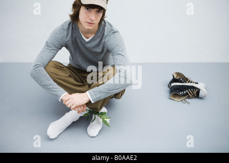 Giovane uomo seduto per terra accanto alle sue scarpe, tenendo fiore, guardando la fotocamera Foto Stock