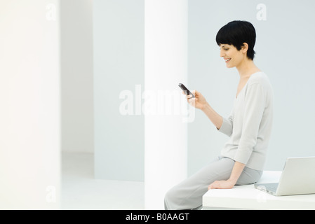 Donna seduta sul bordo della scrivania, guardando il telefono cellulare e sorridente, vista laterale Foto Stock