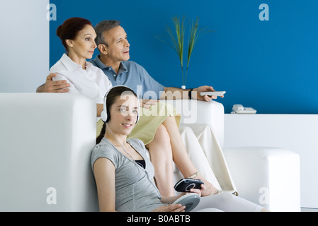 Teen girl ascolto di un lettore CD portatile, sorridente alla fotocamera, i suoi genitori a guardare la TV in background Foto Stock