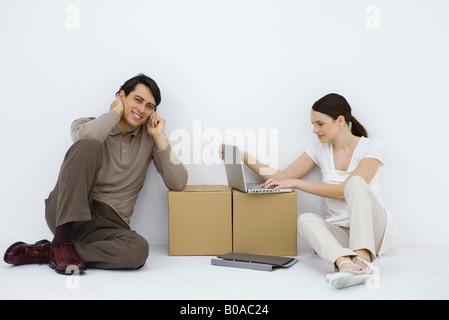 Giovane seduto alla scrivania di fortuna, donna utilizzando il computer portatile, l'uomo tramite telefono cellulare e sorridente in telecamera Foto Stock