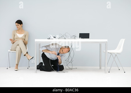 Uomo in ginocchio sotto la scrivania, il collegamento di fili aggrovigliati, donna seduta in cattedra, guardando a portata di mano Foto Stock