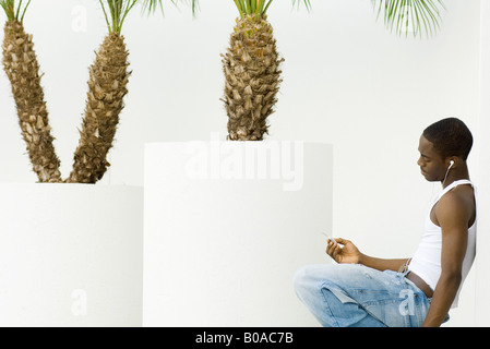 Teen boy ascoltando mp3 player, accovacciato, appoggiata contro il muro, palme in vaso in background Foto Stock