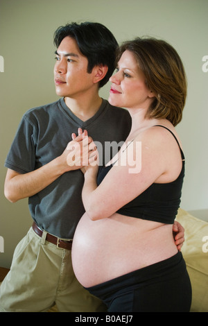 I giovani coniugi uomo asiatico e incinta Donna caucasici in posa insieme nella loro camera da letto modello rilasciato Foto Stock